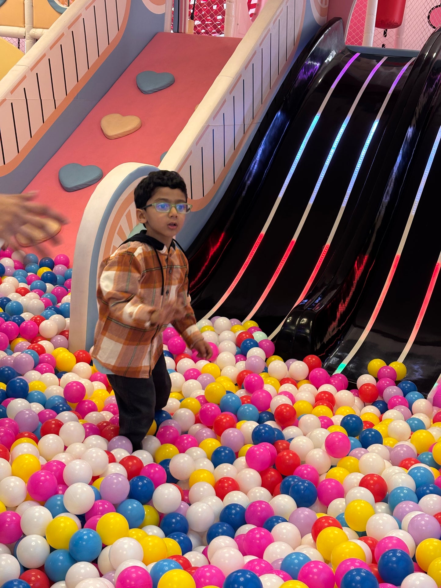 A young boy in glasses stands in a ball pit next to a colorful slide.