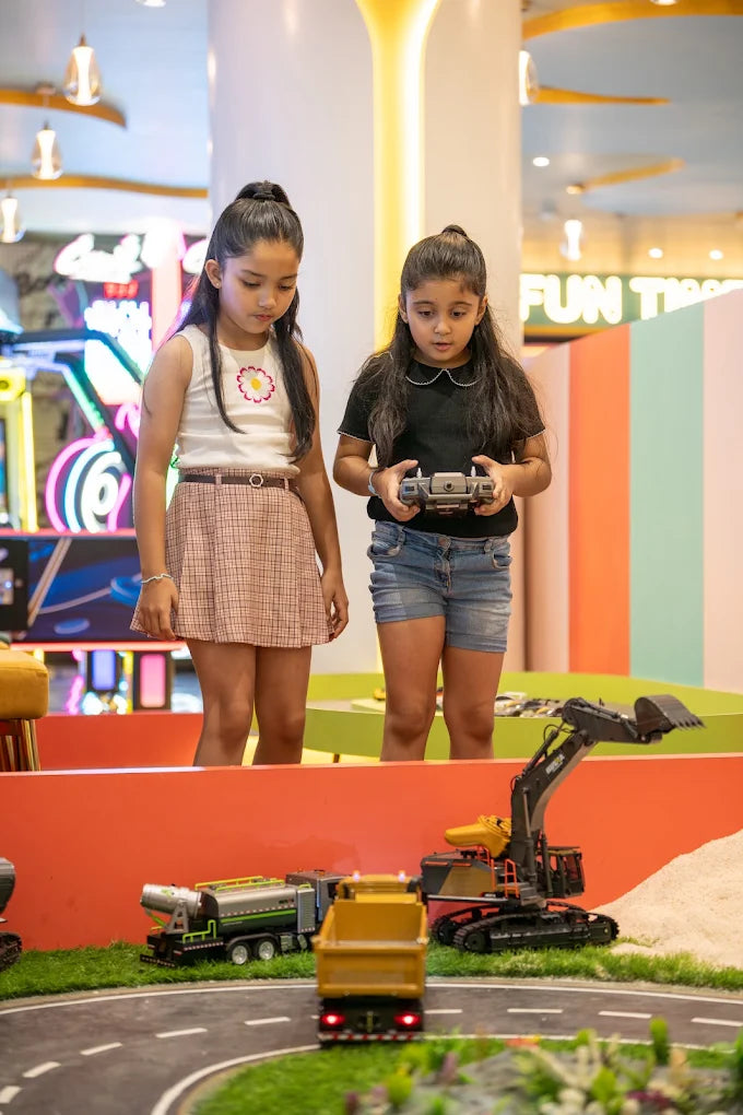 Two young girls play with remote-controlled construction toys in an indoor play area.
