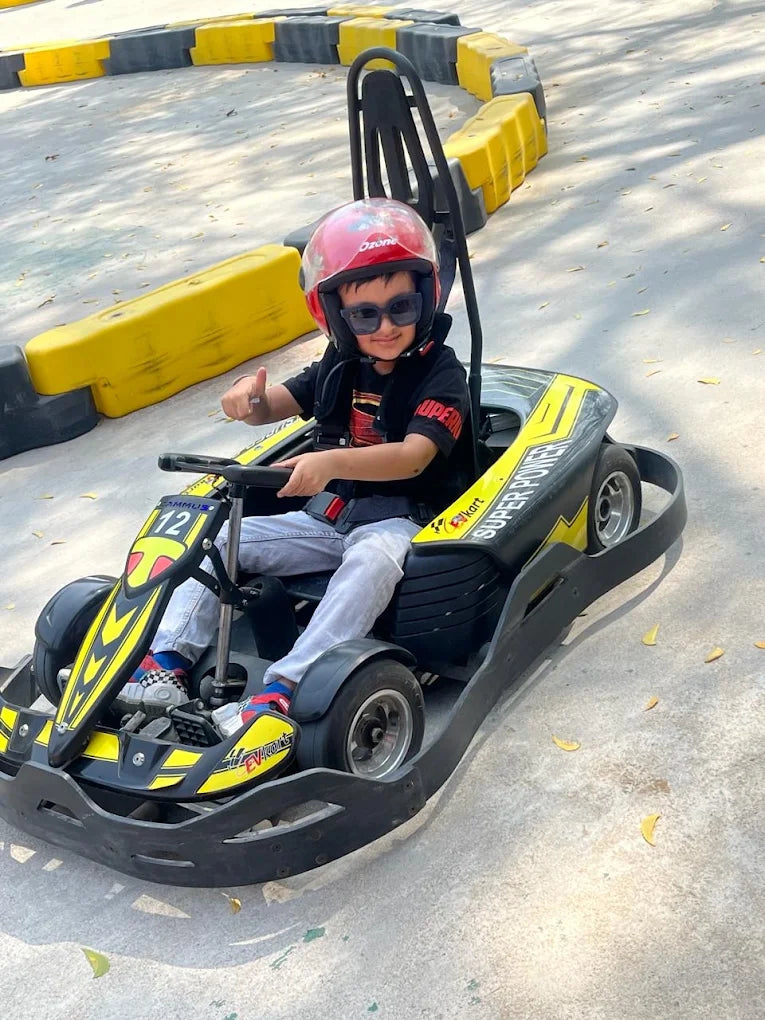 A young boy in a helmet and sunglasses gives a thumbs-up while riding a go-kart on a track.