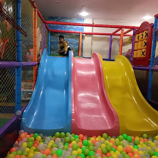 A child sits at the top of a colorful indoor playground slide, ready to slide into a ball pit.