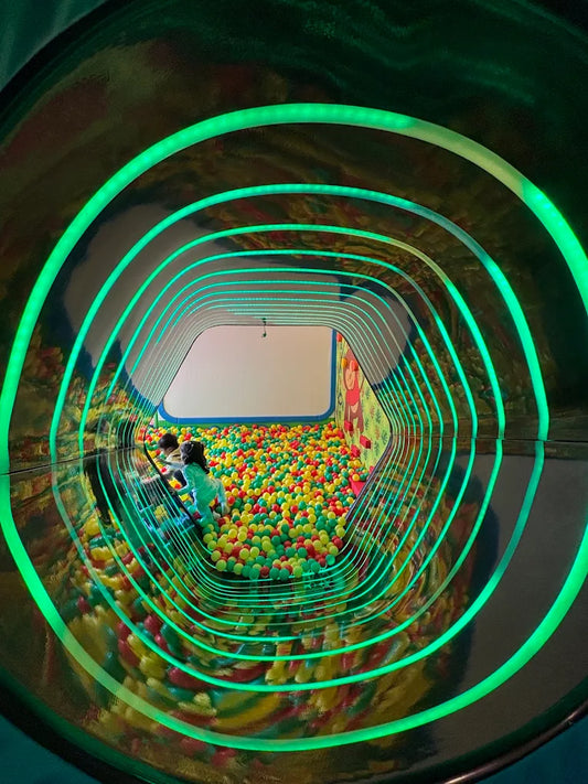 Children play in a ball pit viewed through a tunnel with green neon lights.