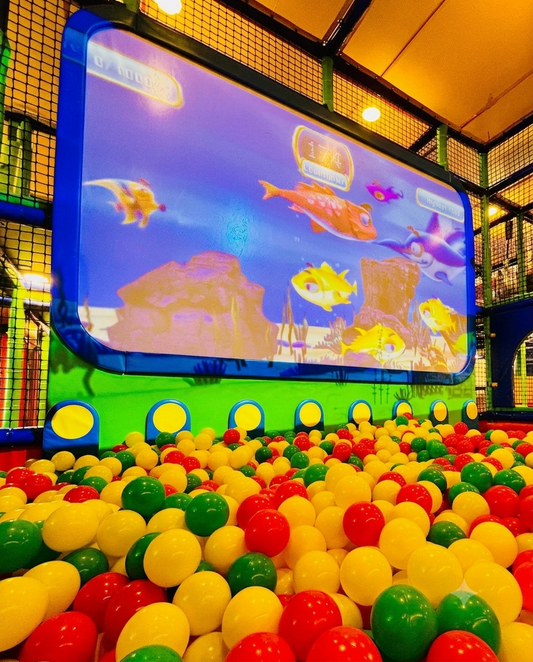 Children play in a ball pit in front of a large screen displaying an underwater scene with animated fish.