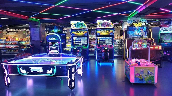 Arcade with neon lights and various game machines, including racing games and an air hockey table.