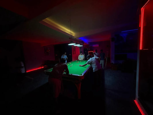 Young men play billiards in a dimly lit room with red and blue neon lighting.