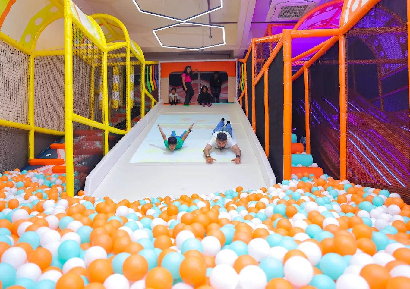 Children and an adult slide down a large white slide into a ball pit in a colorful indoor playground.
