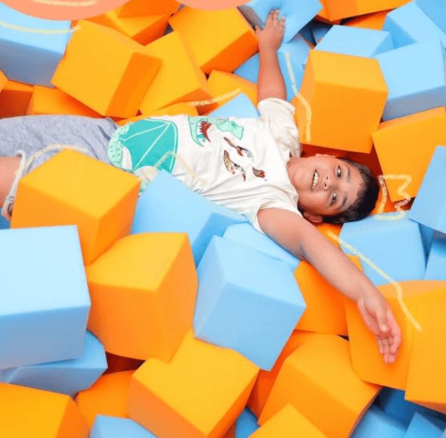 A young boy smiles while lying in a ball pit filled with blue and orange foam cubes.