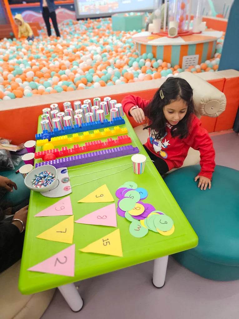 A young girl plays with colorful number cutouts on a green table in a ball pit play area.