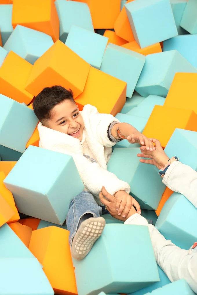A young boy laughs while playing in a ball pit filled with blue and orange foam cubes. Another child's hand reaches out to him.