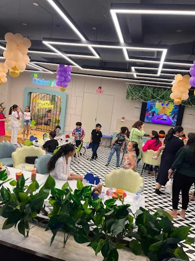Children and adults at a birthday party with balloons and a screen displaying colorful graphics.
