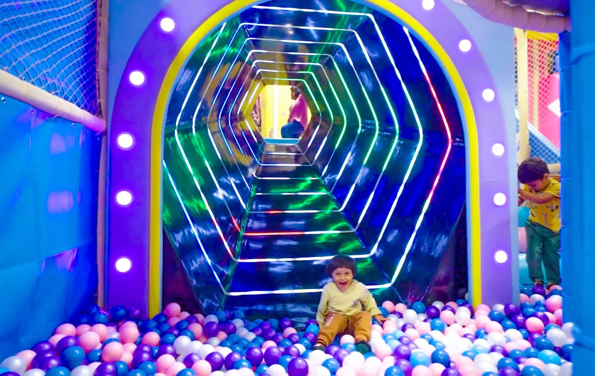 A young child sits in a ball pit in front of a colorful, illuminated tunnel, while another child plays nearby.