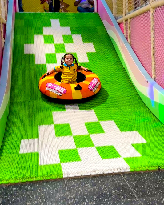 A young child slides down a green and white checkered slide on an inner tube.