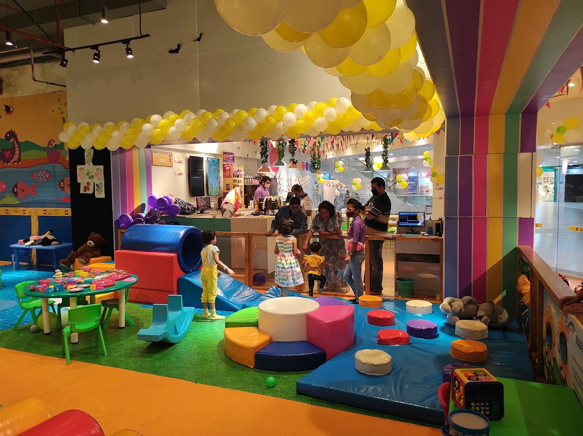 Children play in a colorful indoor playground with balloons and soft play structures. Adults supervise as kids enjoy the fun environment.