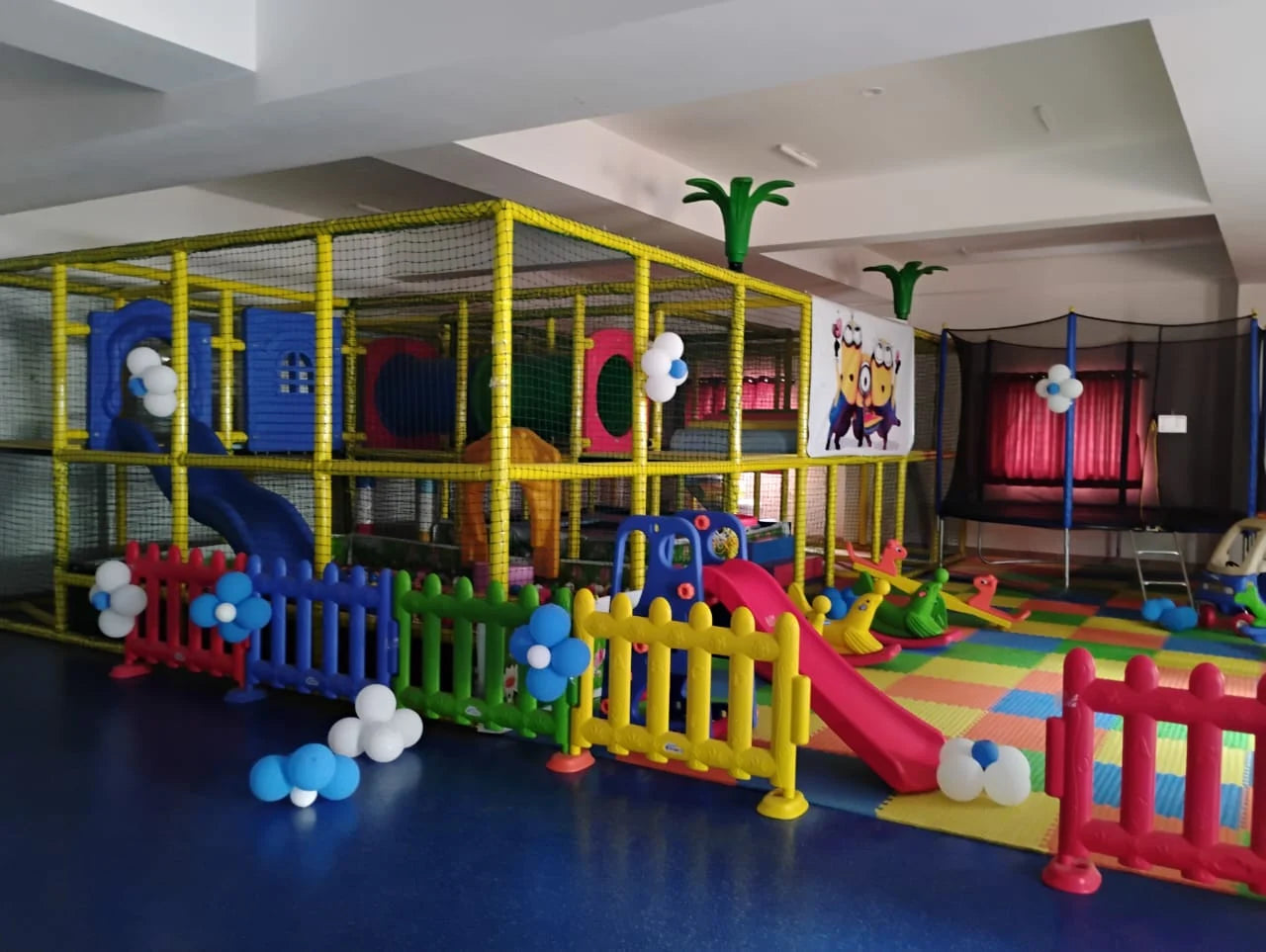 A colorful indoor playground with a large soft play structure, slides, and a trampoline for young children.