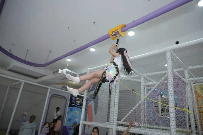 Child wearing safety harness gliding on indoor zipline over play structure at Kidz Funtale Lajpat Nagar event