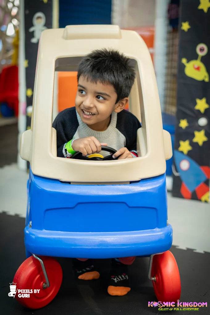 Happy child pretending to drive a blue and beige toy car inside Kosmic Kingdom Sector 66 Gurgaon play area