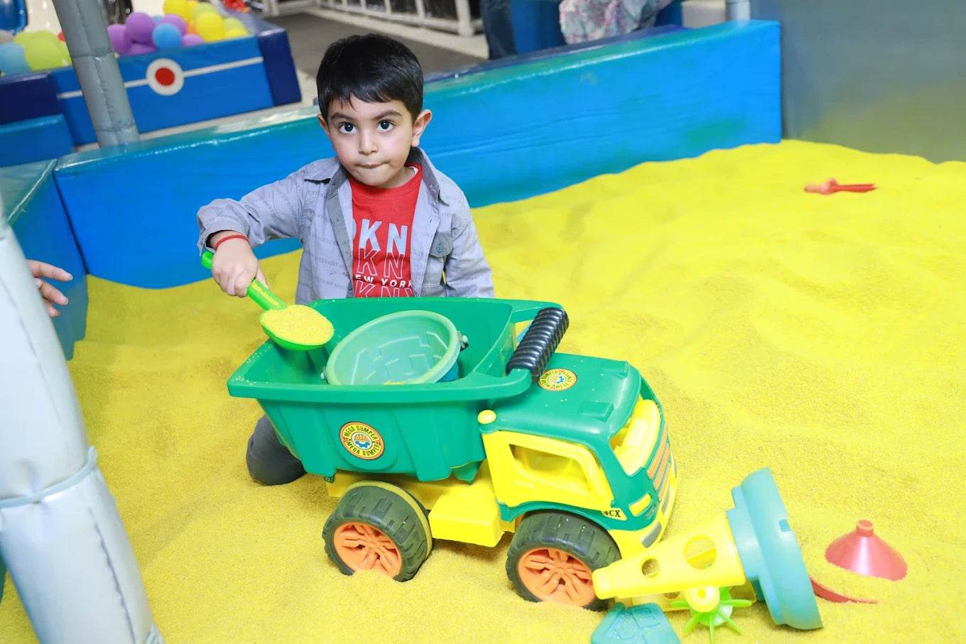 Young boy playing with a green toy dump truck in a yellow sand play area at Kosmic Kingdom Sector 66 Gurgaon