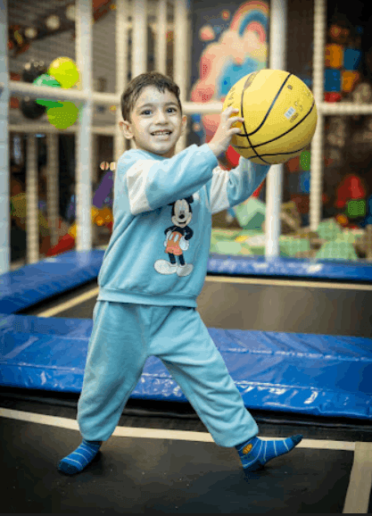 Smiling young boy holding a yellow basketball while playing on a trampoline inside a colorful indoor play area