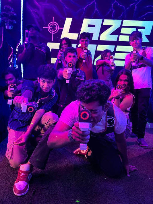 Diverse group aiming laser tag guns posed dynamically in front of purple-lit Lazer Crazer sign at Hauz Khas Delhi location