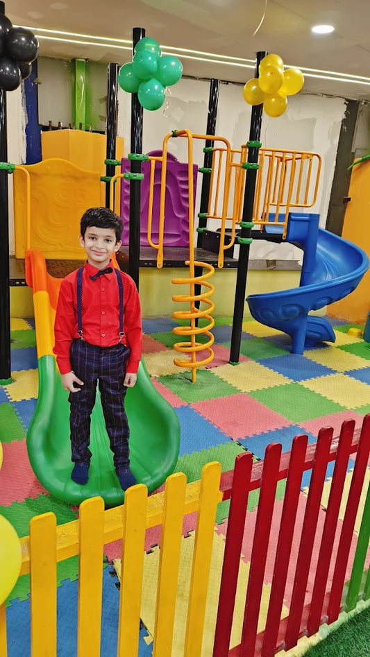 Young boy in formal wear standing on green slide with colorful play structures at Let’s Sunday Play Area Indirapuram Ghaziabad