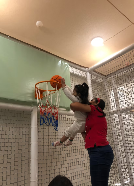 A woman lifts a young child to dunk a basketball into a hoop in a play area.