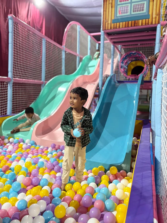 A young boy stands in a ball pit, holding a blue ball, with slides and a play structure behind him.