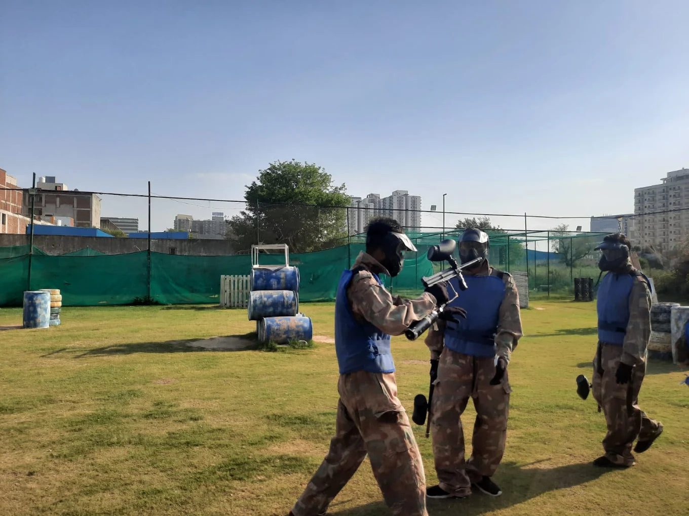 Young adults in camouflage gear and masks play paintball on a grassy field with barrels as obstacles.