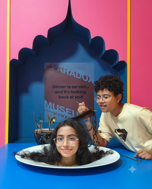 A young person playfully pretends to cut another's hair, who is seated in a large plate, at a museum exhibit.
