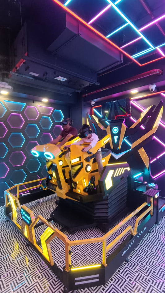 Two people wearing VR headsets ride a yellow futuristic simulator in a neon-lit arcade.