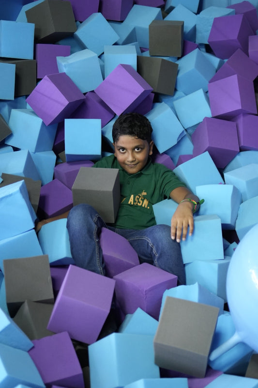 A young boy sits in a ball pit filled with colorful foam cubes. He is wearing a green shirt and blue jeans.