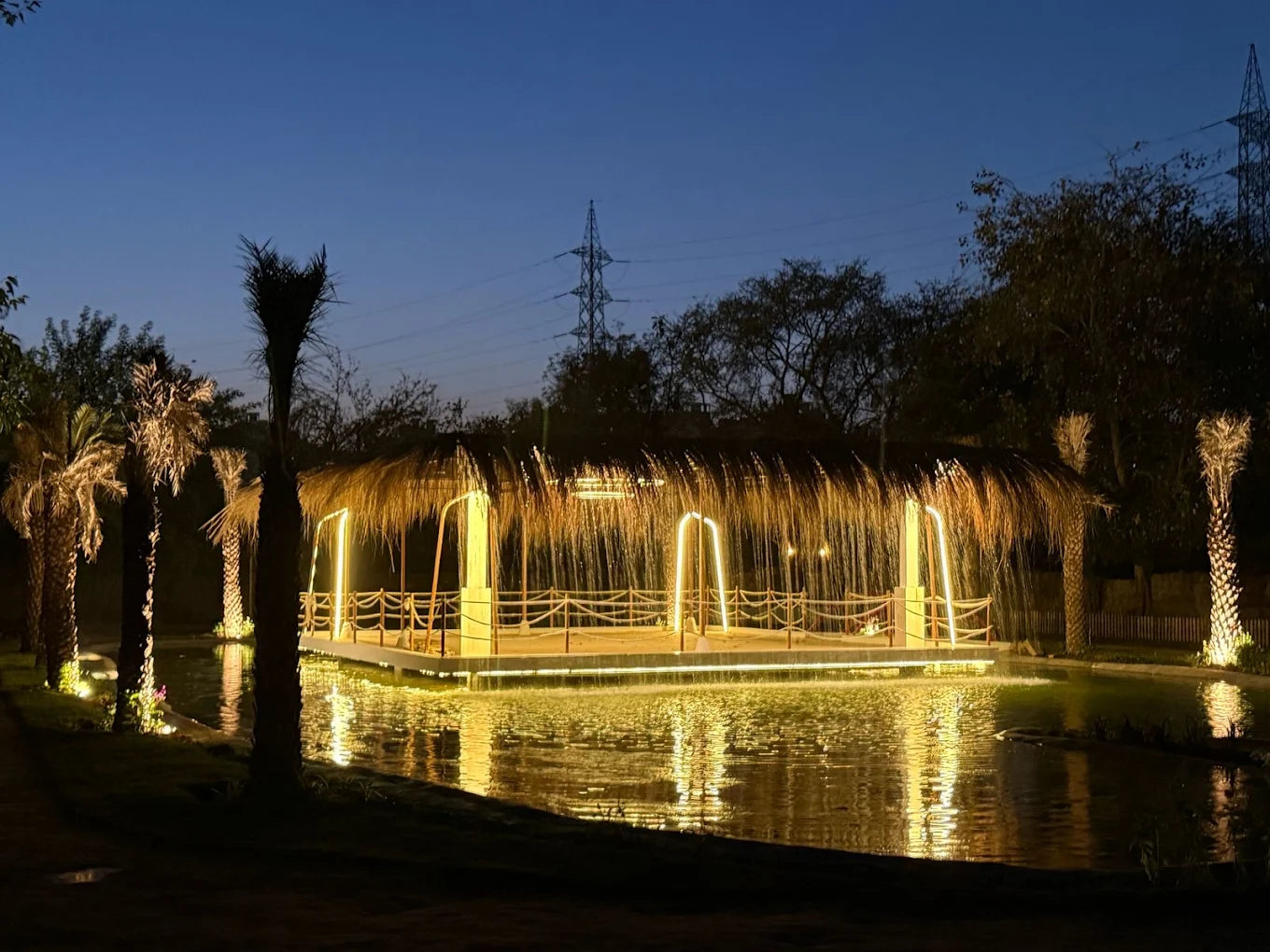 A thatched-roof pavilion with lights sits on a pond at dusk, surrounded by palm trees.