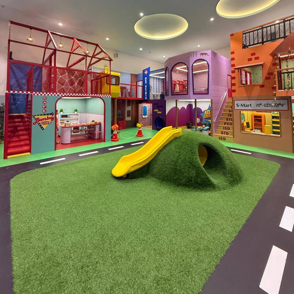 A colorful indoor playground with a yellow slide, a grassy hill, and play structures for children.