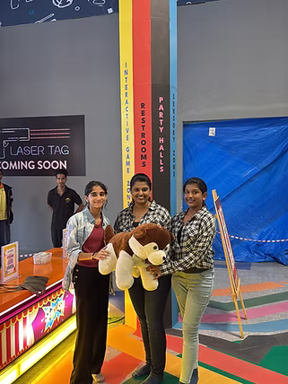 Three young women hold a stuffed dog in front of a laser tag sign.