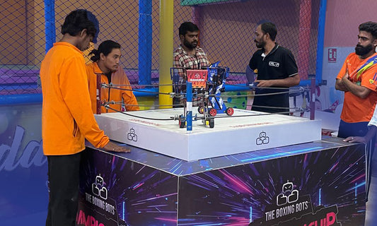 Young adults watch a robot boxing match in an indoor arena.
