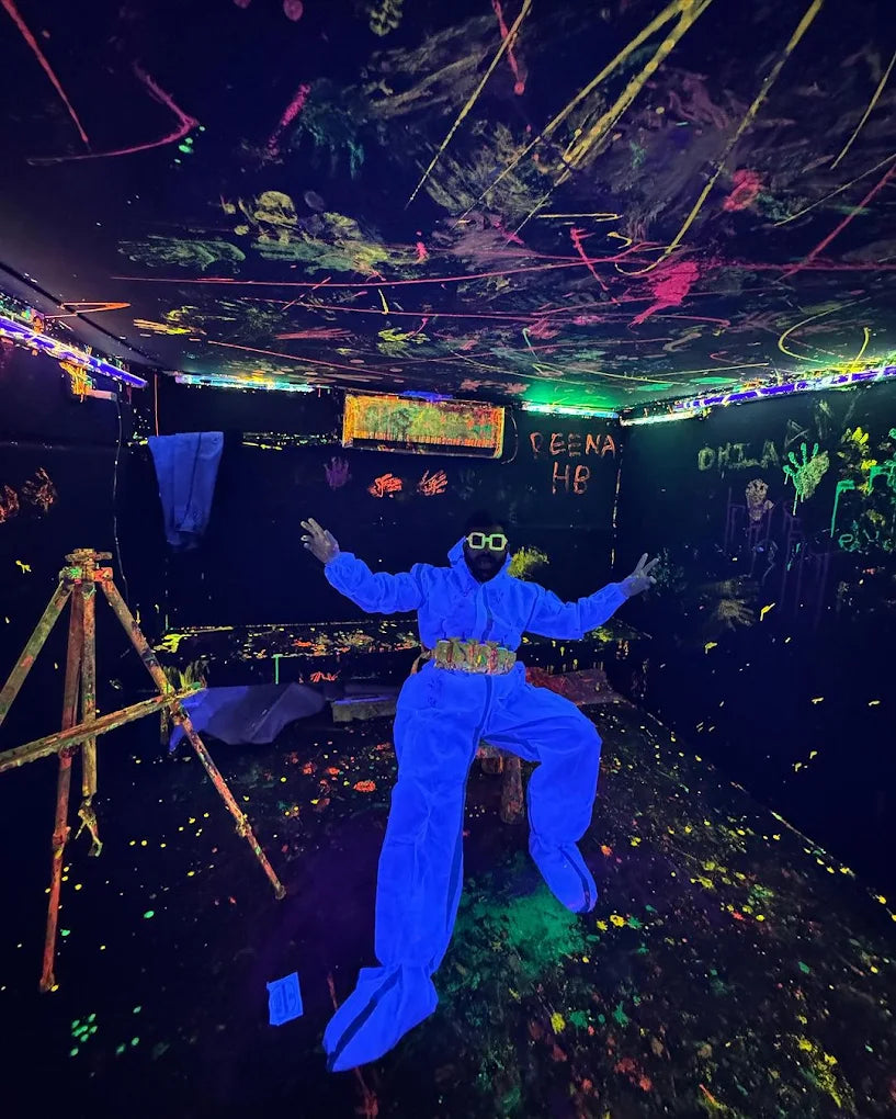 A person in a blue suit and goggles poses in a room covered in neon paint splatters under blacklights.
