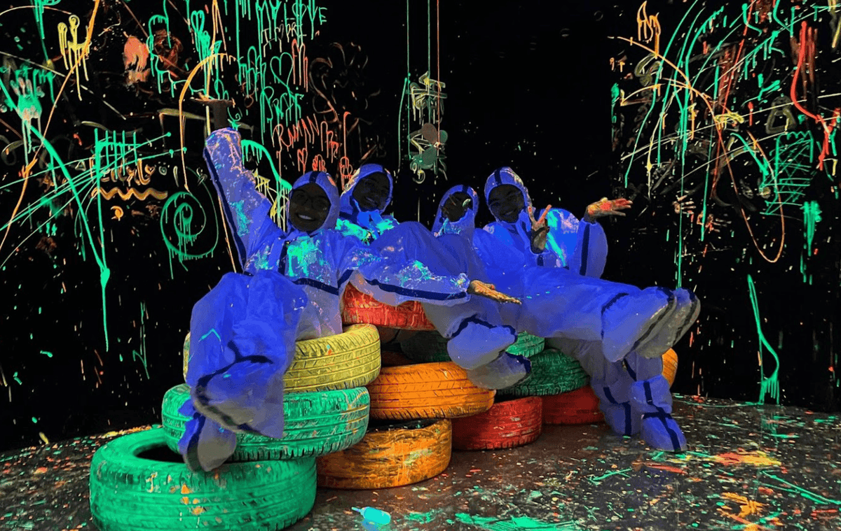 Four young people in neon paint-splattered suits pose on colorful tires in a blacklight room.