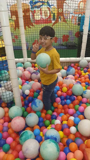 A young boy in a yellow shirt plays in a ball pit, holding a light blue ball and making a peace sign.
