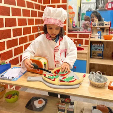 Child playing with toy pizza and utensils in a pretend kitchen setting.