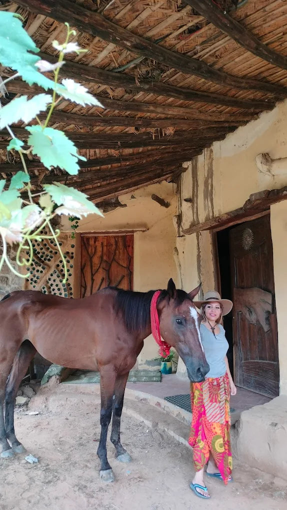 A woman in a hat and colorful pants stands next to a brown horse in front of a rustic building.