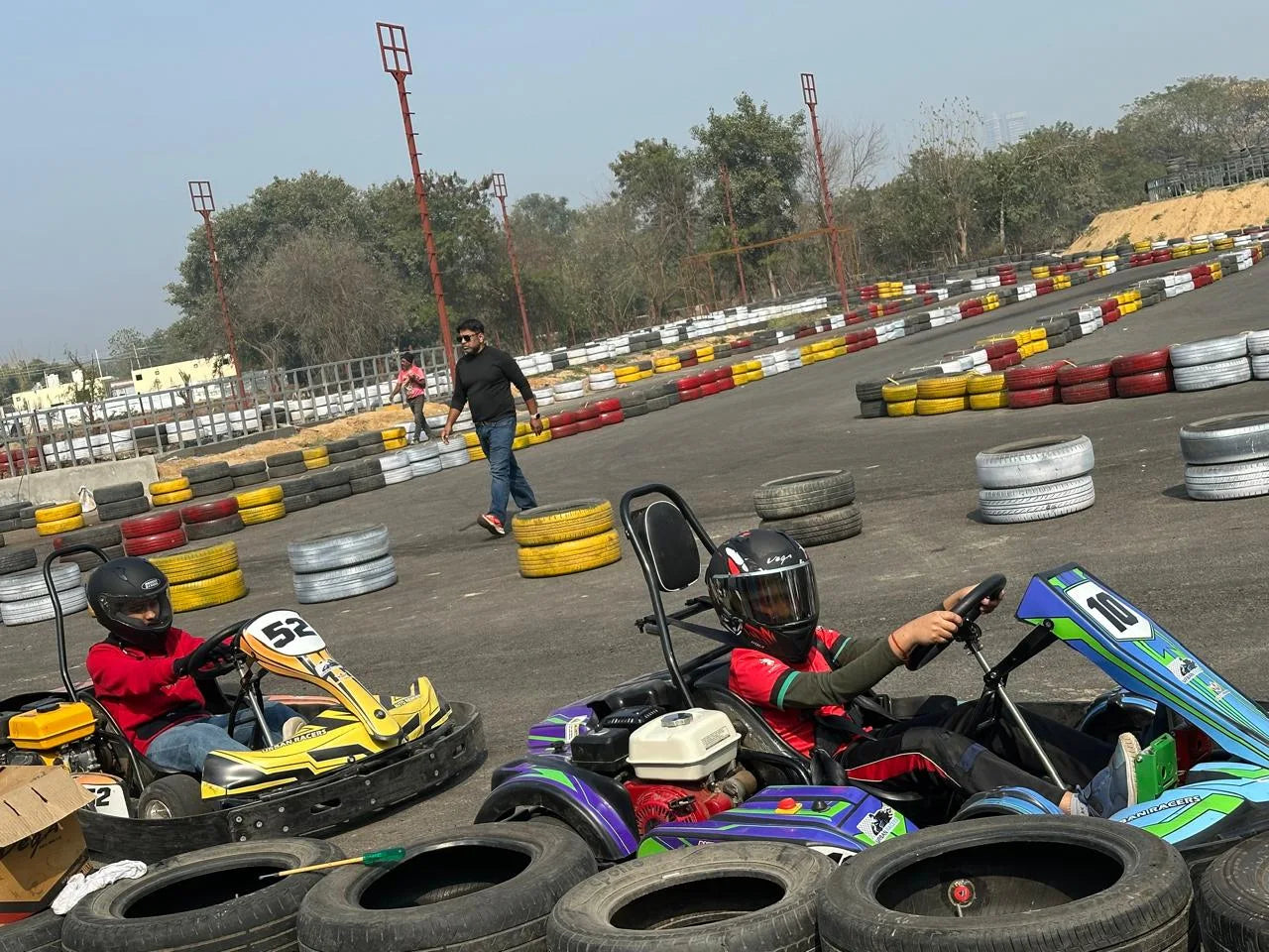 Two children race go-karts on a track, with a man walking in the background.