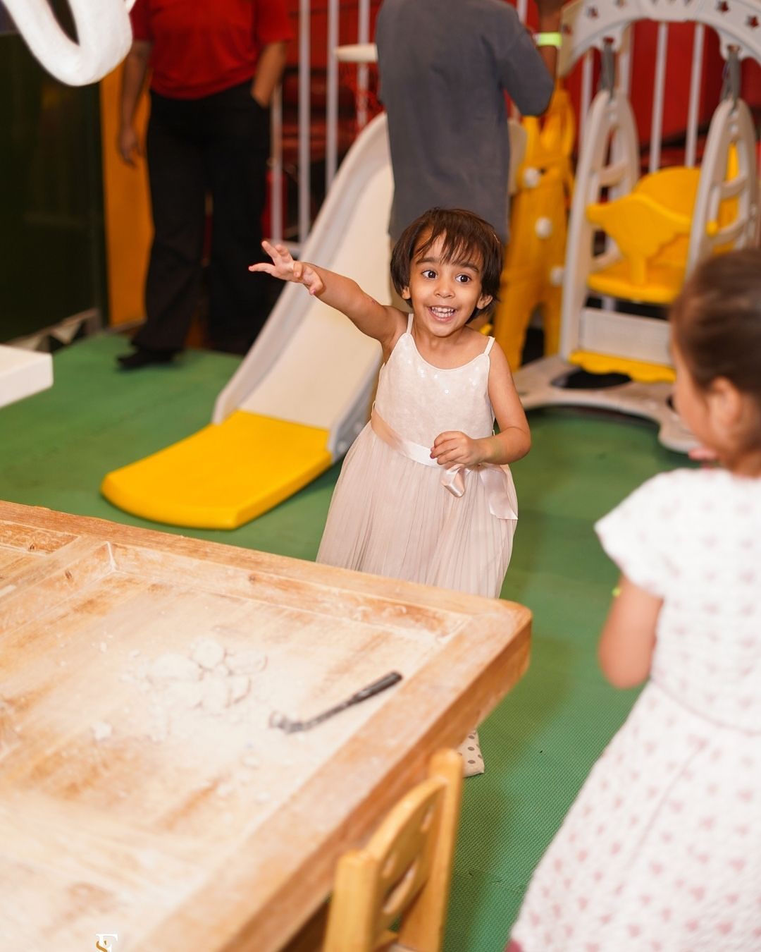 A young girl in a light pink dress smiles and reaches out in a play area. Another child is visible in the foreground.