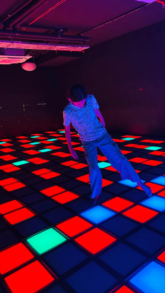 A child dances on a colorful, illuminated disco floor. The floor has red, blue, and green glowing squares.