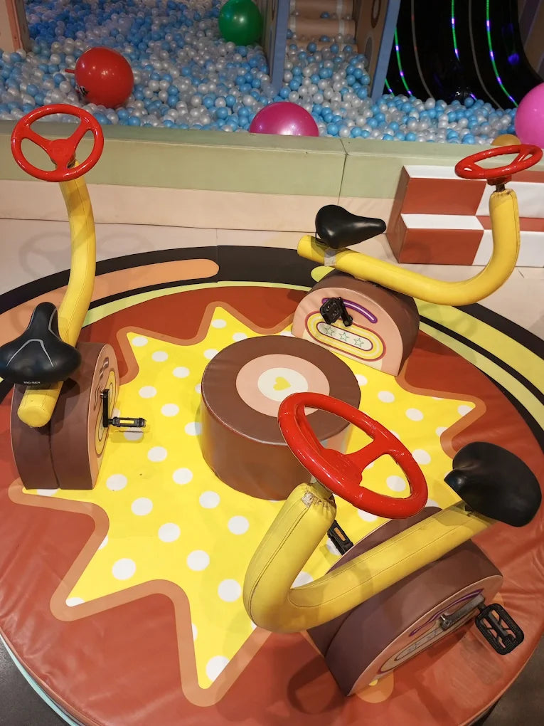 Interactive pedal ride with red steering wheels on a bright patterned floor near a blue ball pit at indoor activity center