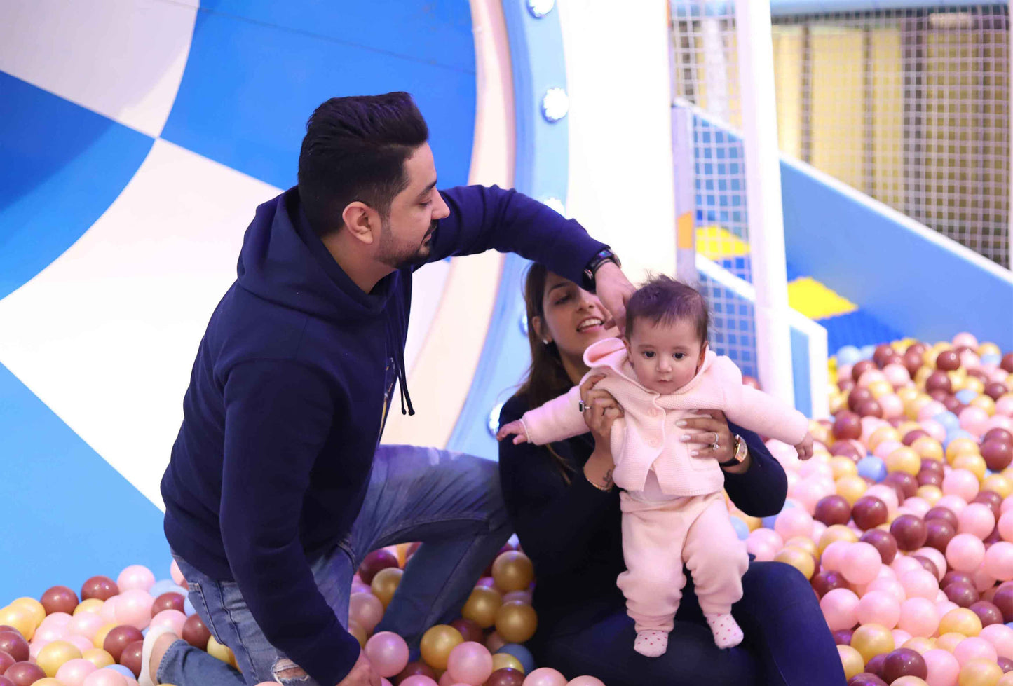 Parents playing with their baby in a soft ball pit area with colorful balls and padded play structures nearby