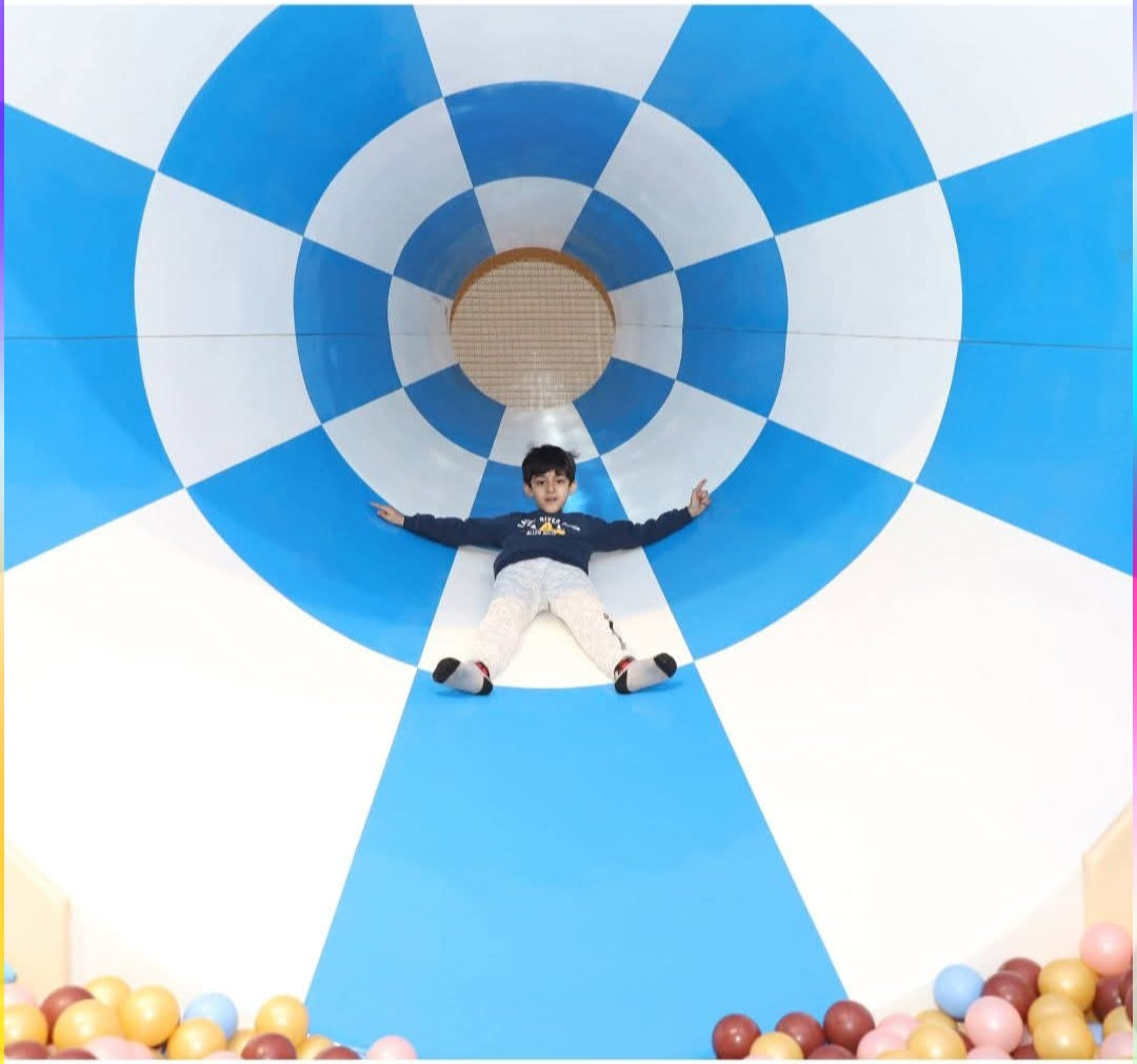 Child sliding down a blue and white tunnel slide into a ball pit with pastel-colored balls at Zig Zag Zoh Faridabad venue