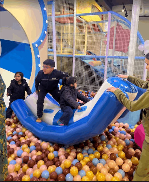 Children playing on a blue inflatable rocking toy surrounded by multicolored balls in an indoor activity center