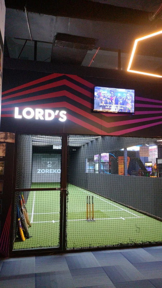 Indoor cricket nets with a "Lord's" sign and a screen showing a crowd.
