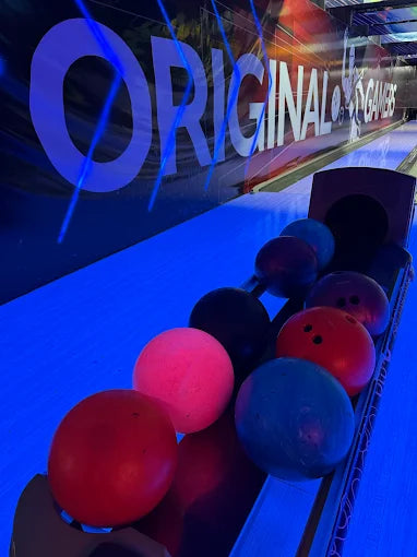 Bowling balls in various colors lined up on rack with illuminated bowling lane and bold wall text in background