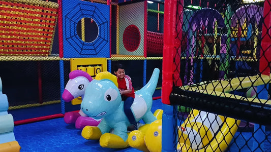 A young child rides a blue inflatable dinosaur in a colorful indoor playground.