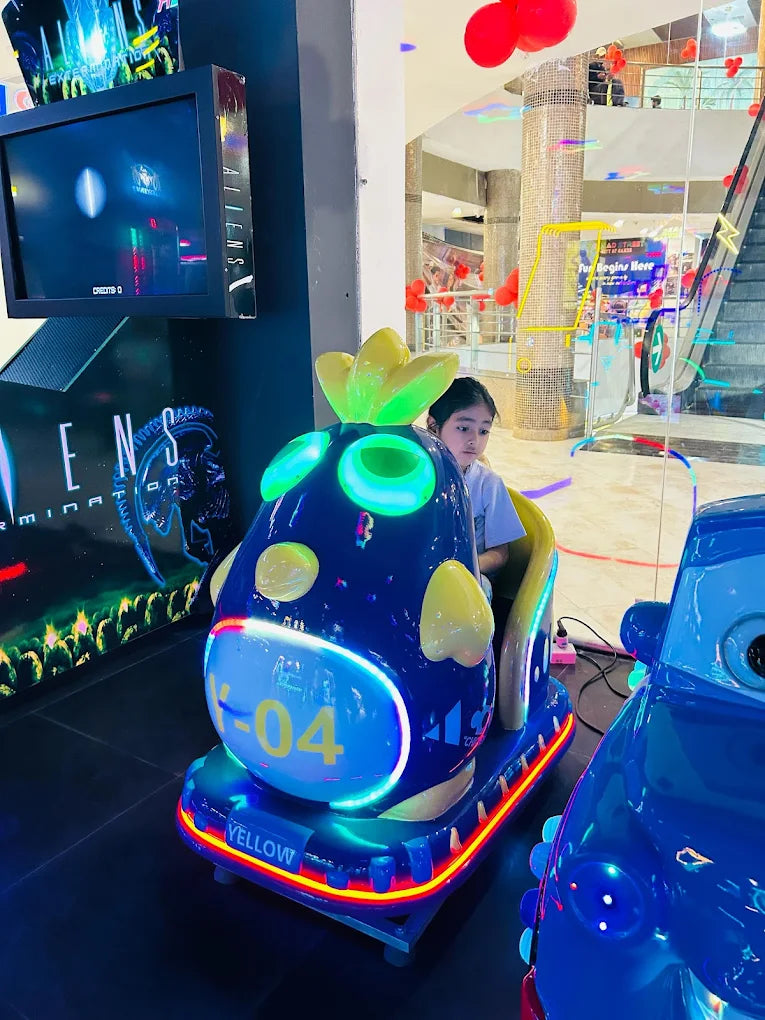 A young girl rides a blue cartoon-themed amusement park ride in a brightly lit indoor arcade.