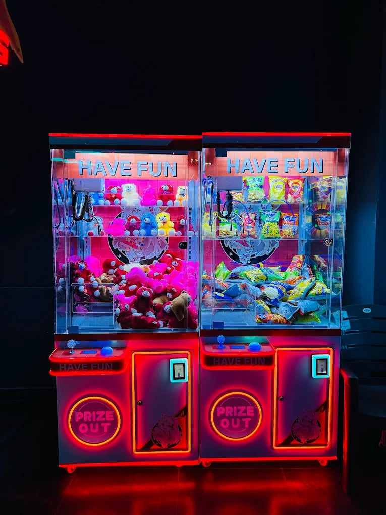 Two brightly lit arcade claw machines filled with plush toys and snacks, inviting players to "Have Fun."